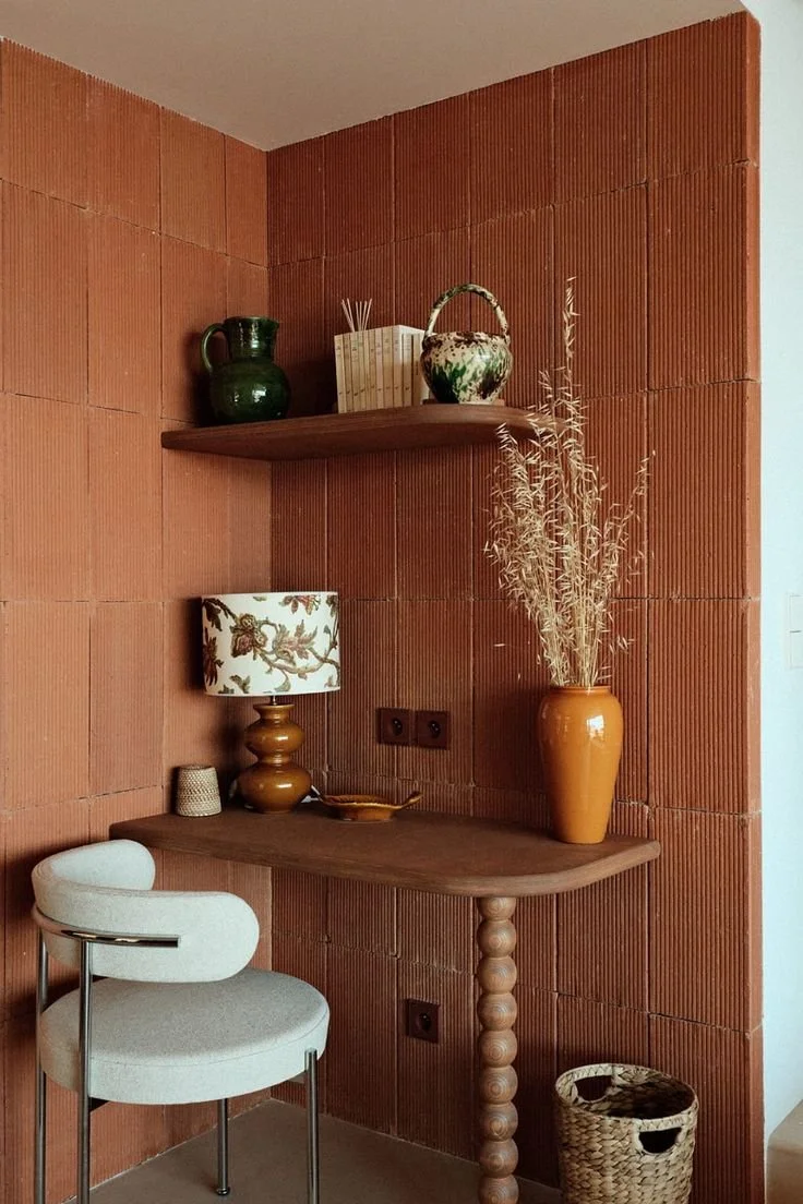 Coin salon avec mur en briques rouges, étagères en bois avec des vases, une lampe de table, une grande vase avec des branches séchées, un fauteuil blanc avec bras en chrome, panier en osier sur le sol.
