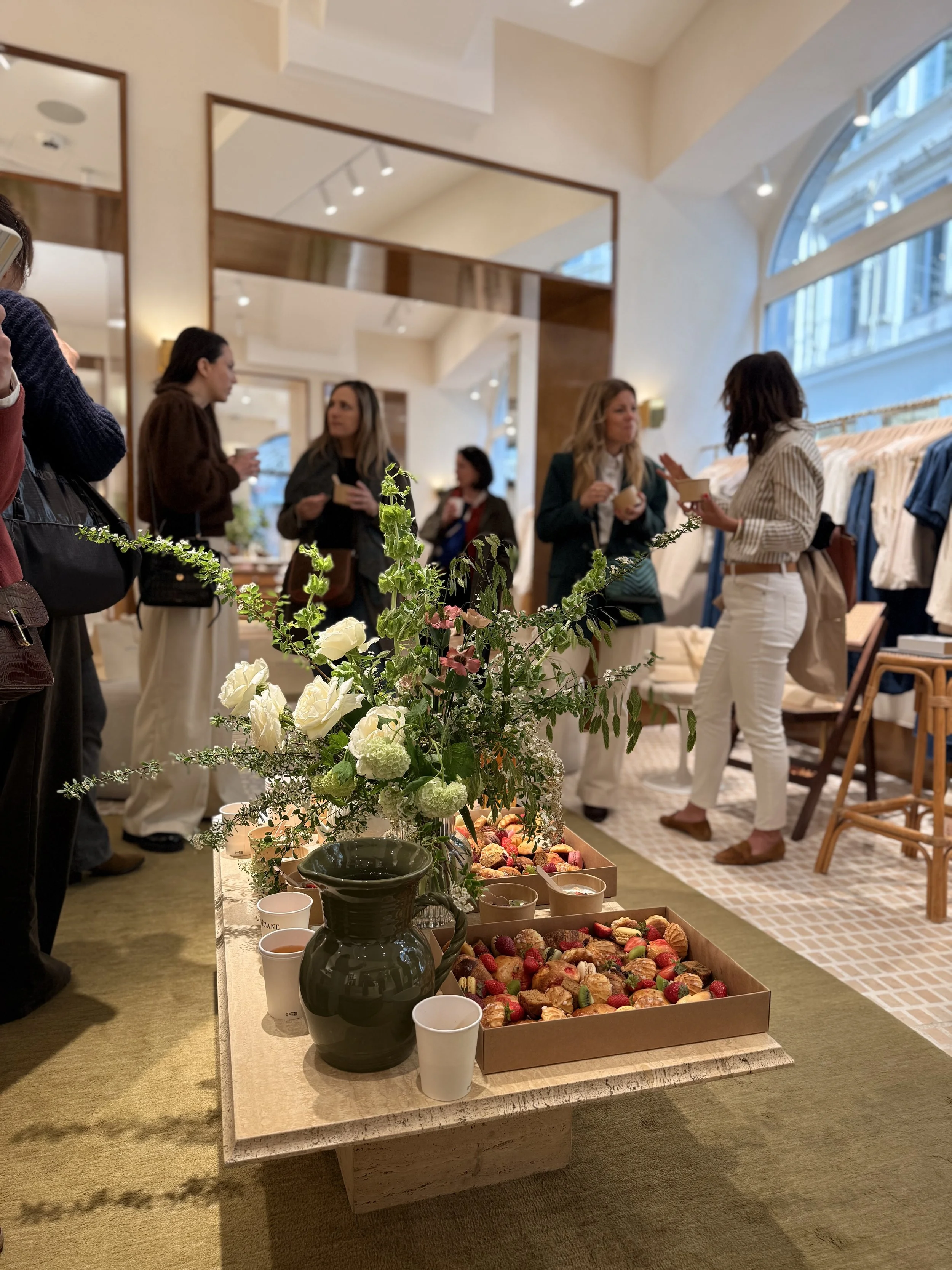 Un tableau avec un vase de fleurs et un étalage de petits gâteaux et fruits décoratifs dans un intérieur lumineux avec plusieurs personnes discutant.