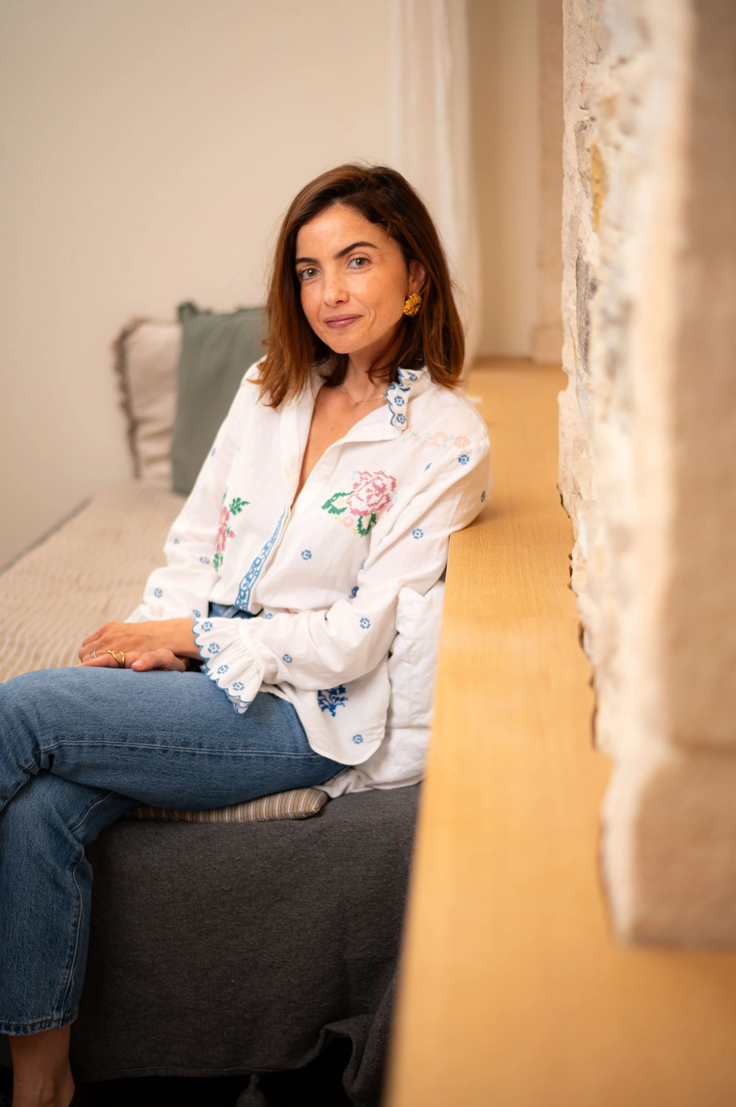 Une femme rousse assise sur un canapé, portant une chemise blanche avec des broderies colorées et des jeans, souriant légèrement, dans une pièce chaleureuse avec un mur en pierre et des coussins derrière elle.