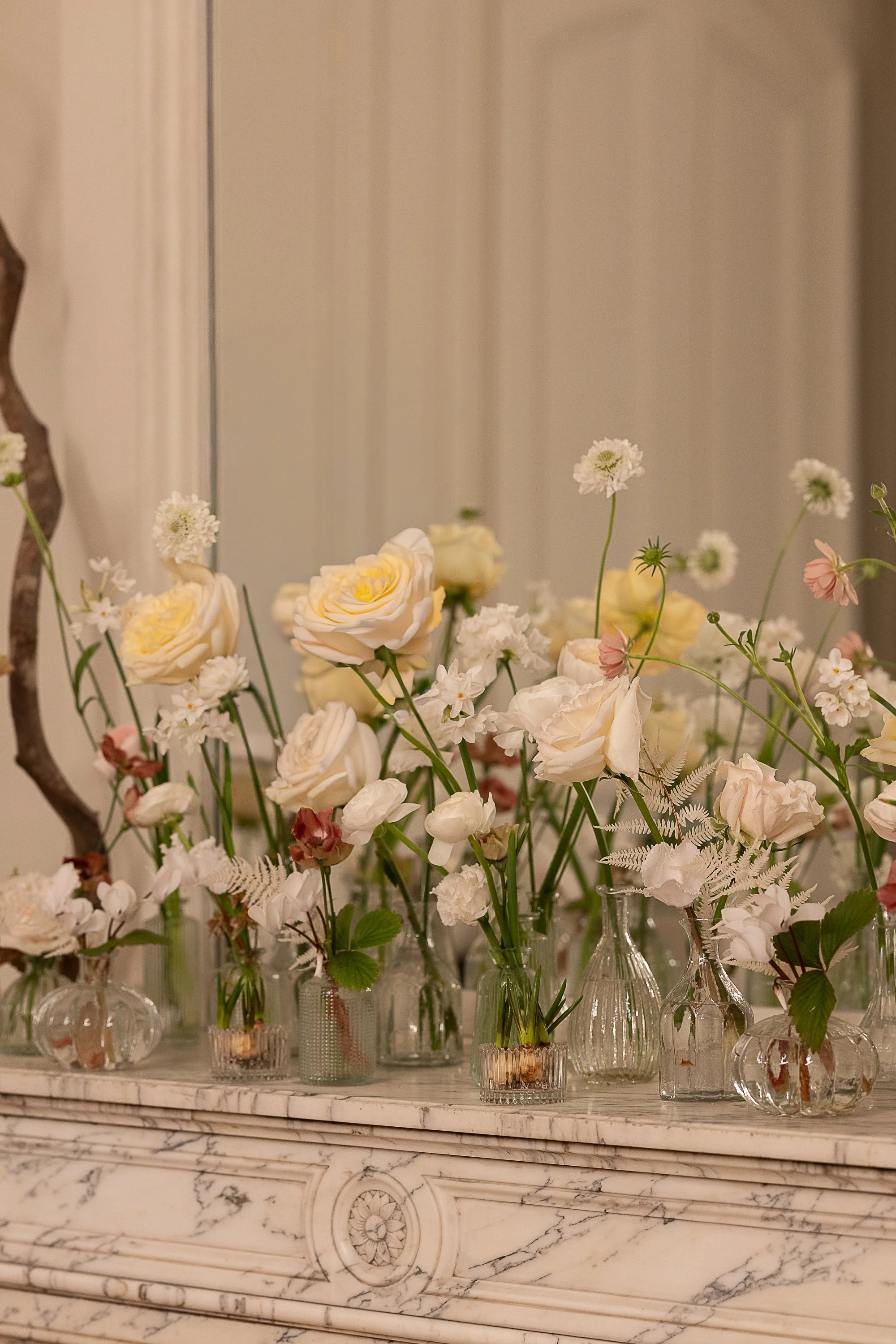 Arrangement floral avec différentes fleurs blanches, roses crèmes et roses pâles dans des petits vases en verre, posés sur une cheminée en marbre blanc avec motifs sculptés.