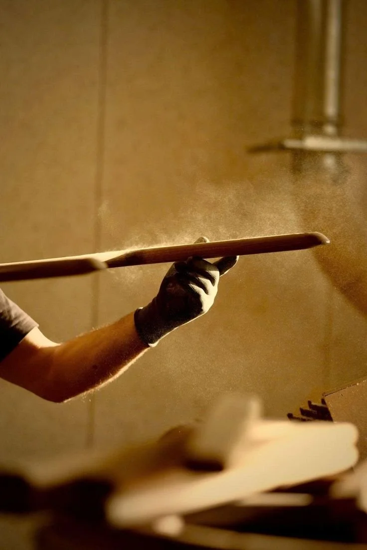 Une main portant un gant noir tient une spatule en bois dans une ambiance chaleureuse, probablement dans une cuisine ou un atelier de fabrication.