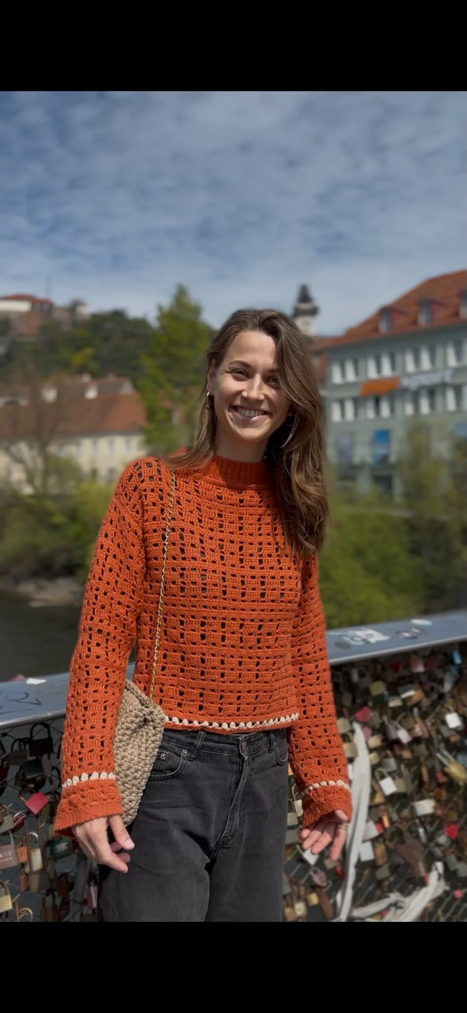 A smiling woman with brown hair wearing an orange crocheted sweater with white accents, black jeans, holding a beige crocheted purse, standing on a bridge with numerous love locks, with buildings, trees, and a cloudy sky in the background.