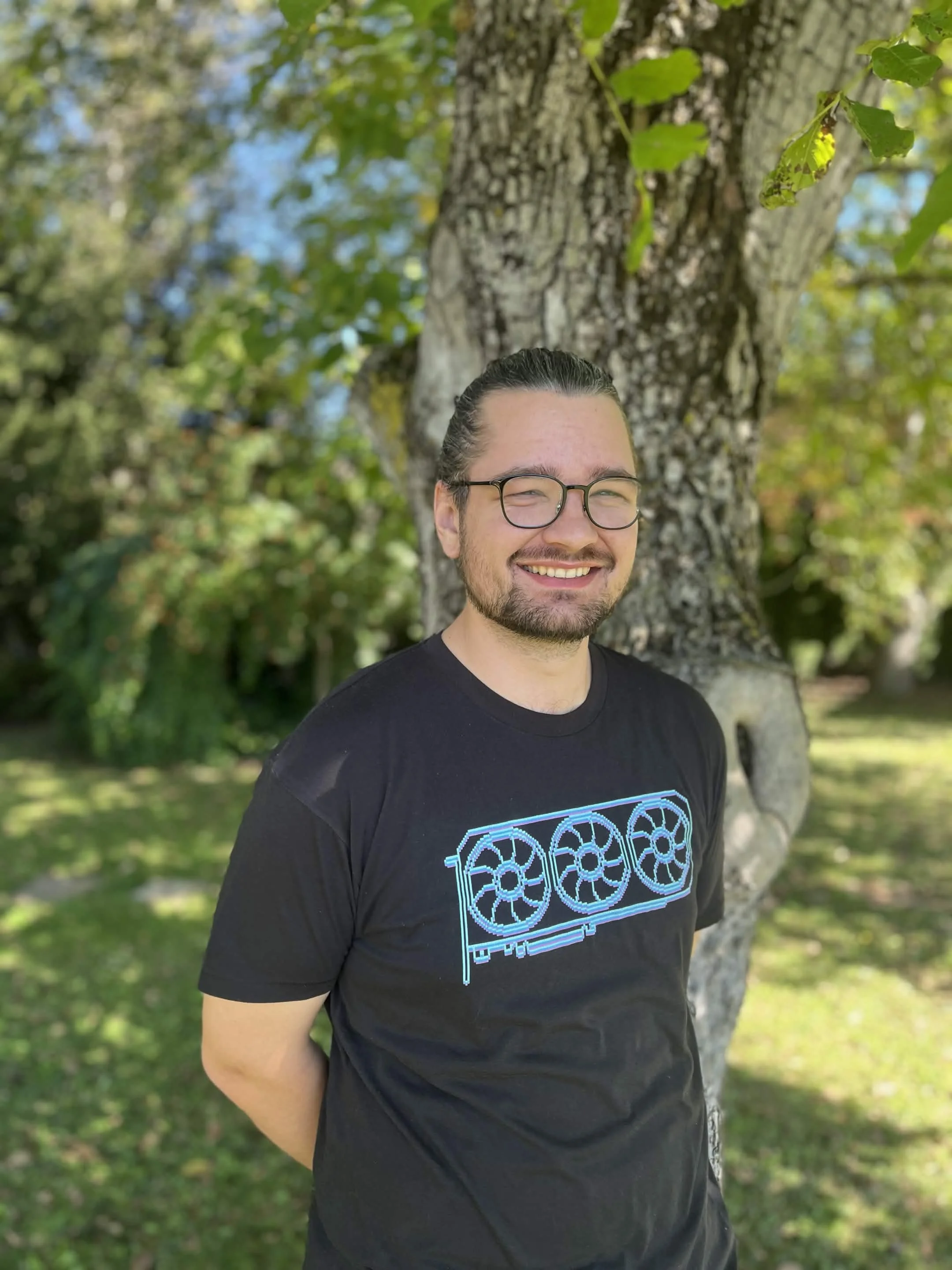 A man with glasses smiling outdoors in front of a tree with green leaves and a blue sky.
