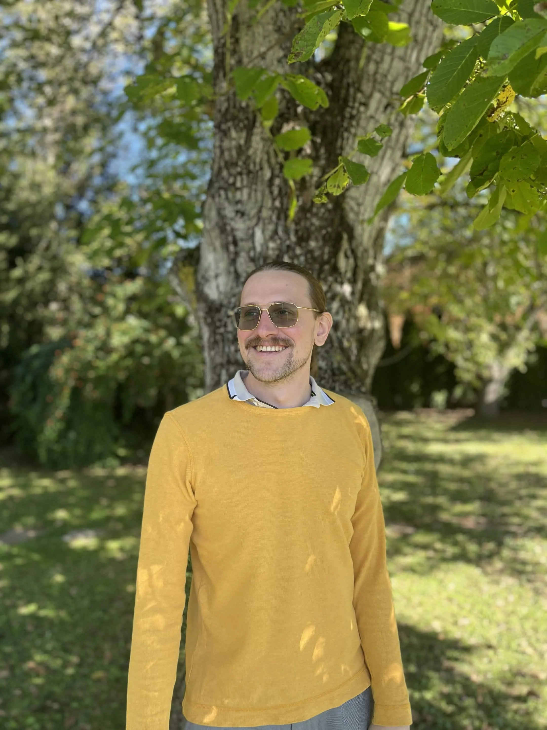 A smiling man with sunglasses, wearing a yellow sweater, standing outdoors in front of a large tree with green leaves and a background of greenery and blue sky.