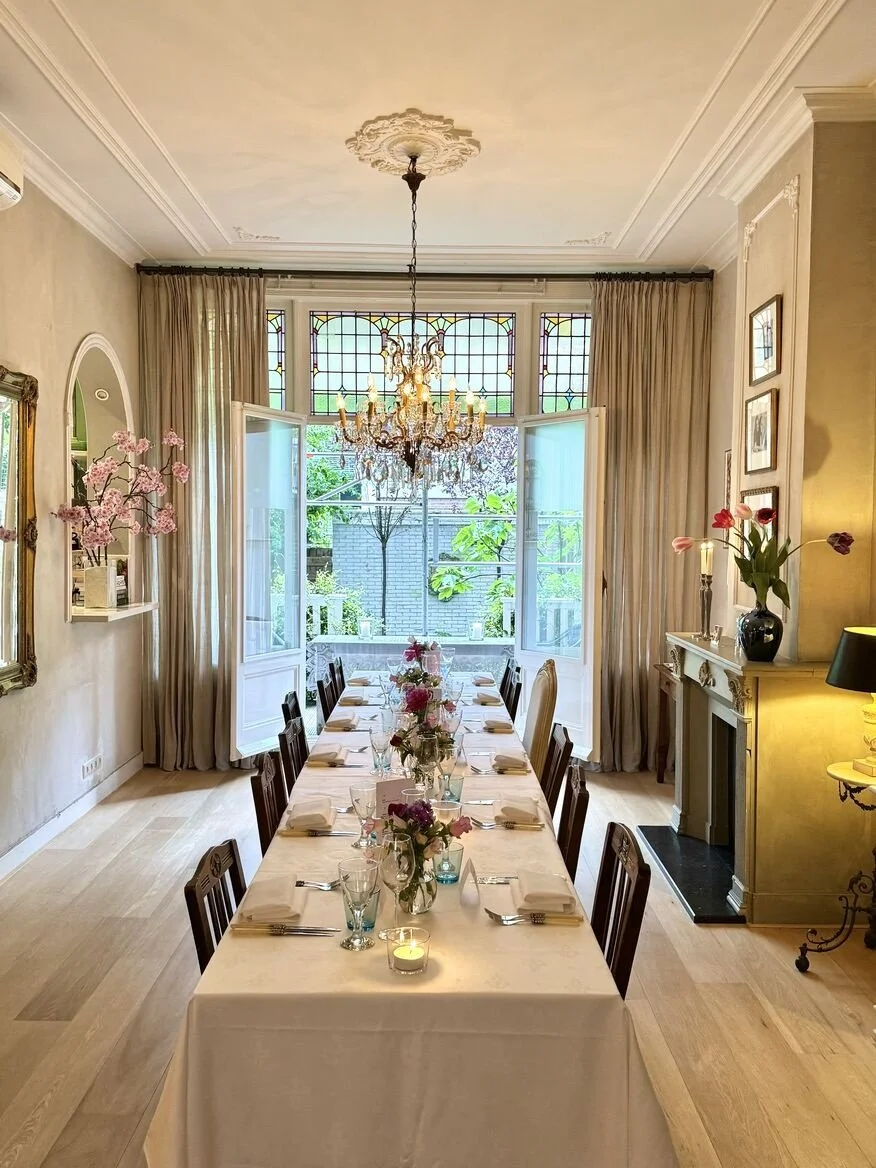 Elegant private dining room in Amsterdam Oud Zuid with a long beautifully set table, white linen, crystal chandelier, fireplace and open French doors overlooking a garden terrace.