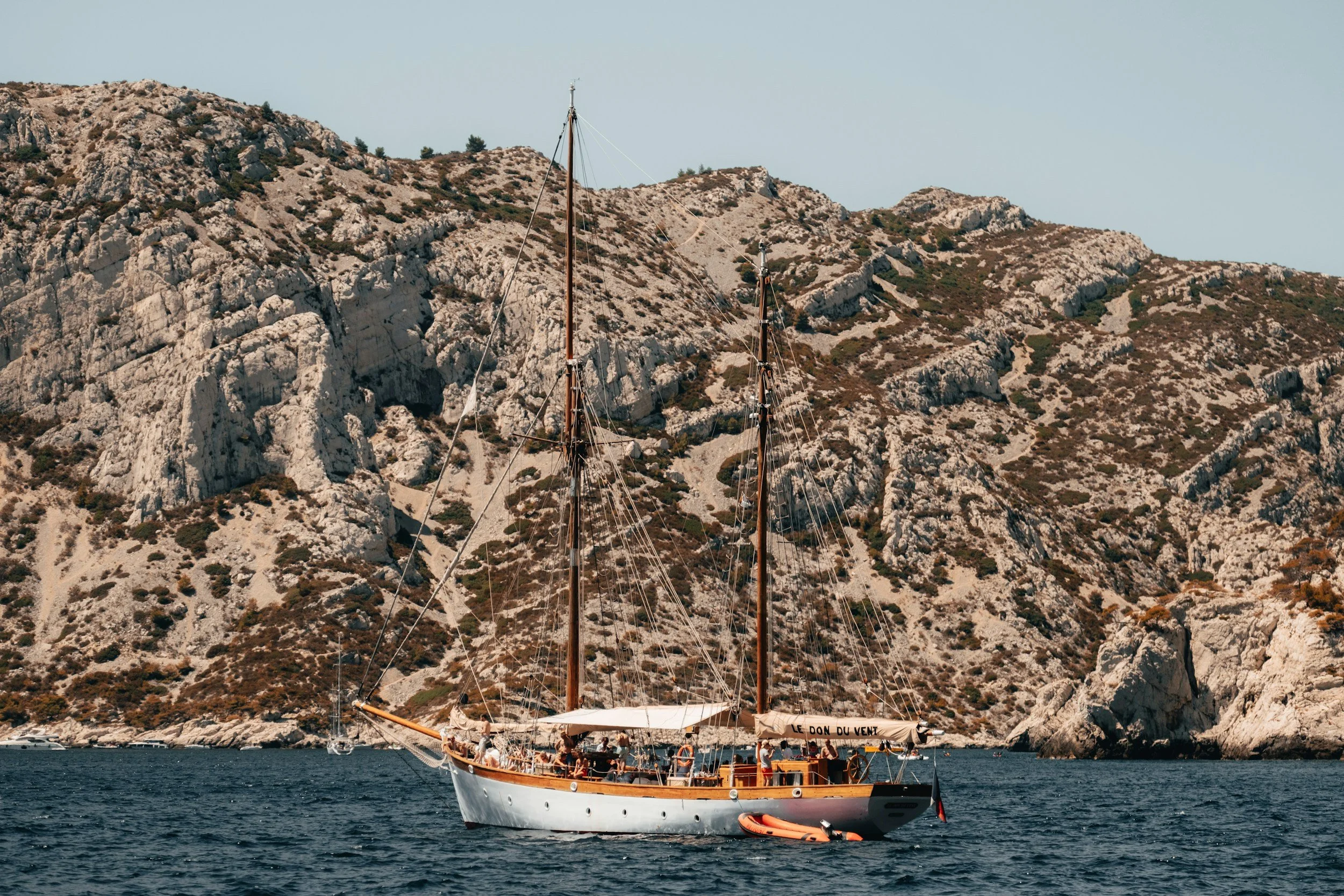 A sailboat with two tall masts is on calm water near a rocky, arid coastline with sparse vegetation.