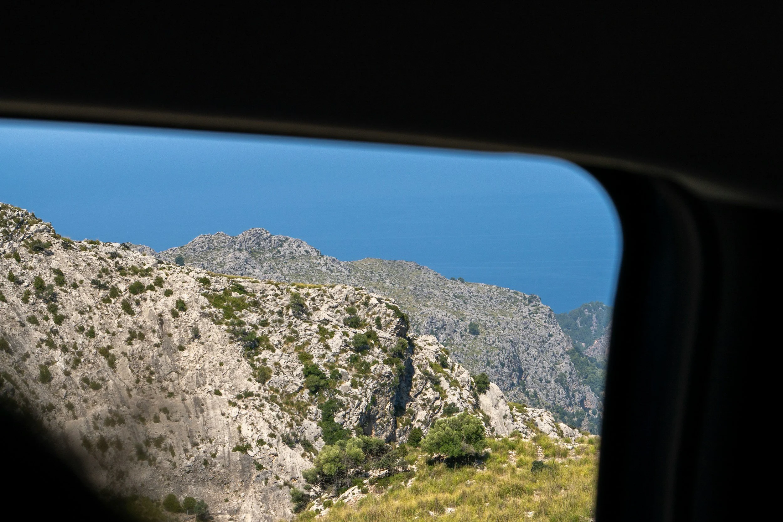 Mountain landscape seen through a vehicle window with rocky hills, green vegetation, and a blue sky in the background.