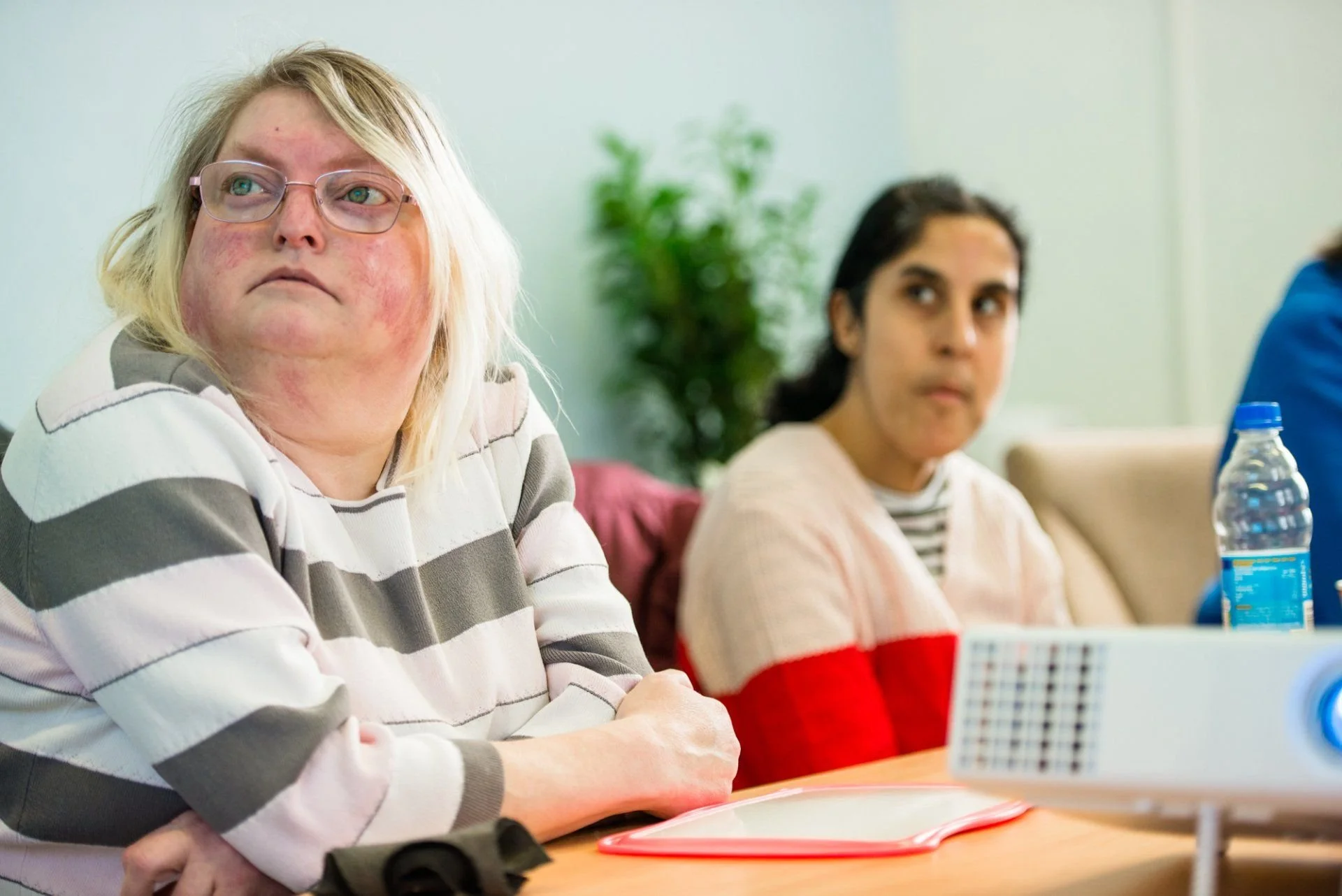 Two women sitting at a table in a meeting room, with a projector in the foreground. The woman on the left has blonde hair, glasses, and a striped sweater, looking to her left. The woman on the right has dark hair and is wearing a white sweater with red sleeves.
