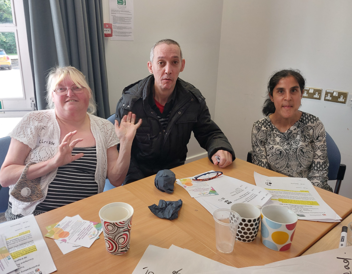 Three people sitting at a table with papers and cups, indoors with a window showing a  carpark outside.
