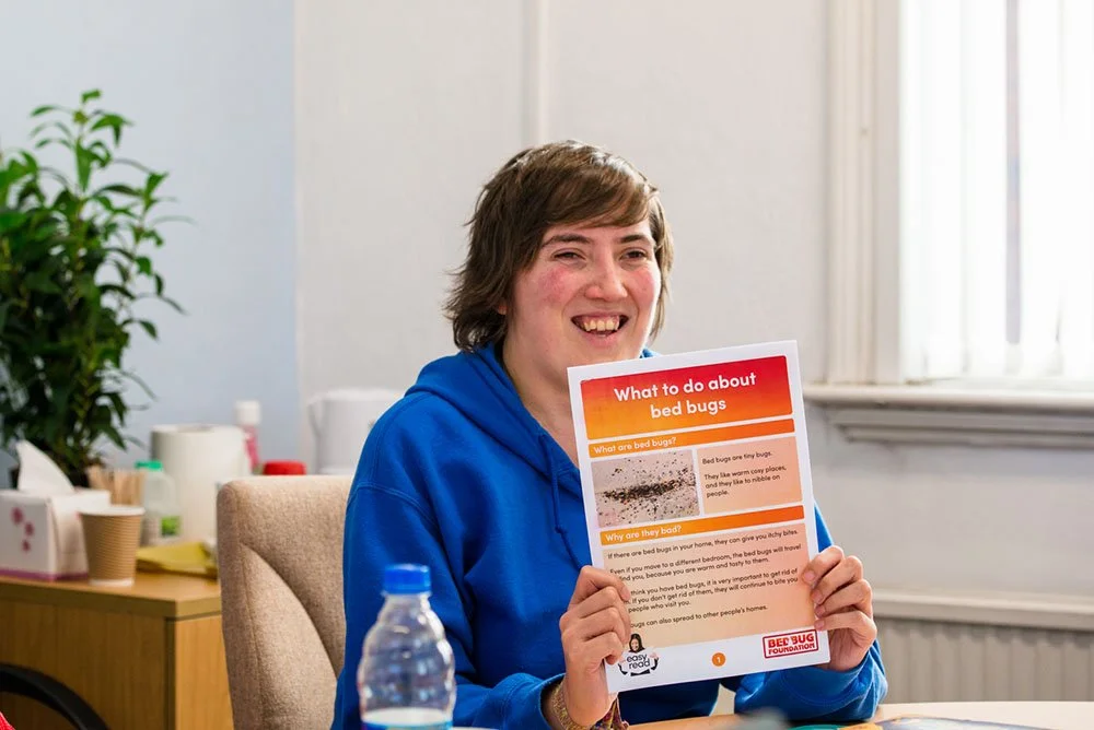 Woman sitting at a table holding a colorful informational flyer about bed bugs, smiling. Office setting with plants, cups, and supplies in the background.