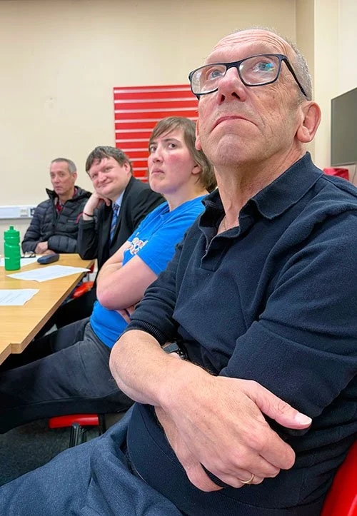 Four people sitting at a conference table in a meeting room, listening attentively. The man in the foreground wears glasses and a black shirt, with arms crossed. The others include a teenage boy in a blue T-shirt and two adults, one with a black jacket and the other with a dark blazer. There are papers, a green water bottle, and electronic devices on the table, with a red-striped wall in the background.