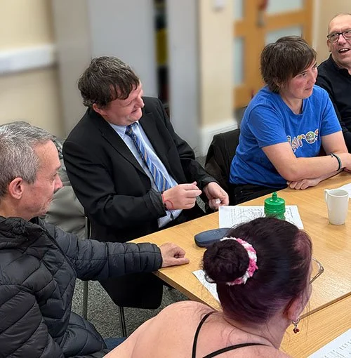 Group of five people sitting around a table during a meeting or discussion, with papers and drinks on the table.