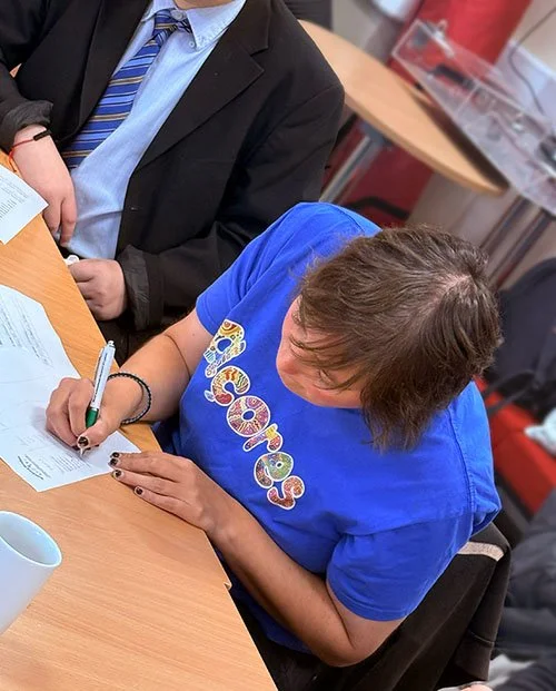 A woman with short brown hair wearing a blue t-shirt, sitting at a table and signing a document with a green pen. A man in a suit and striped tie is visible beside her, and there are papers and a white mug on the table.