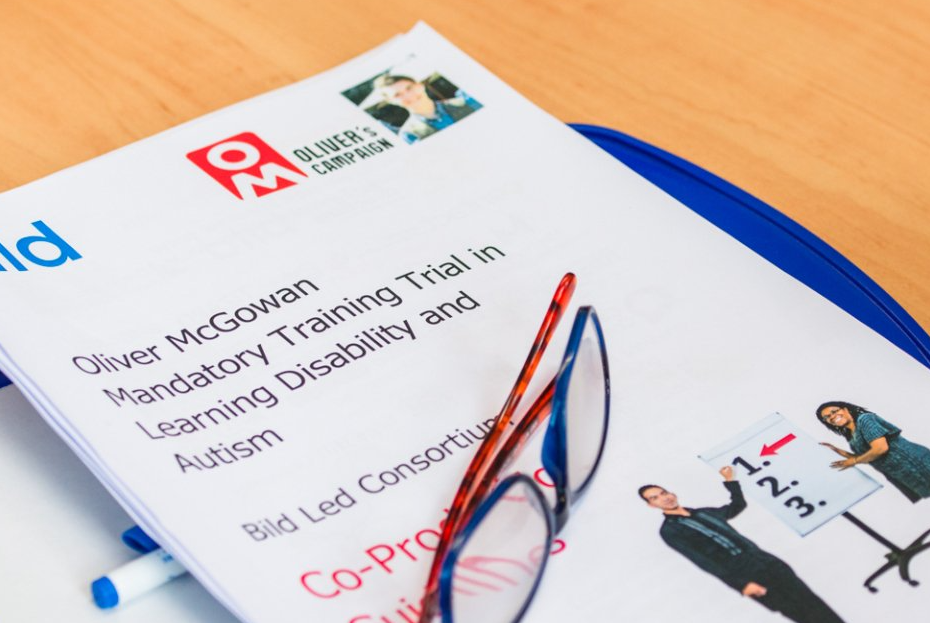 A printed document titled 'Oliver McGowan Mandatory Training Trial in Learning Disability and Autism' with a photo of a person at the top and a small image of two people, one pointing at a whiteboard, on the bottom right. Glasses and a pen are resting on the document.