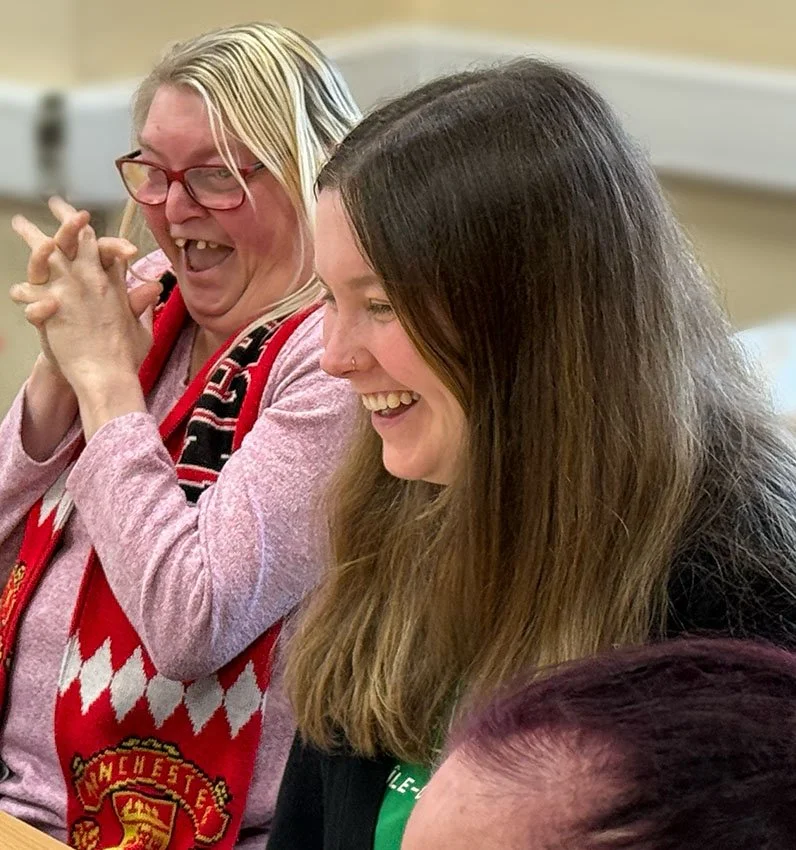 Two women laughing together, one with blonde hair and glasses wearing a pink sweater and Manchester United scarf, the other with long brown hair and a nose ring, in a social setting.