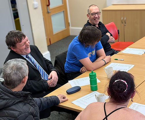 Four people are sitting at a conference table, laughing and talking. There are papers, a water bottle, and cups on the table.