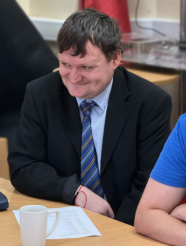 A man with short, wavy brown hair wearing a black suit, light blue shirt, and striped tie sitting at a table in a meeting or office setting, smiling and looking down.