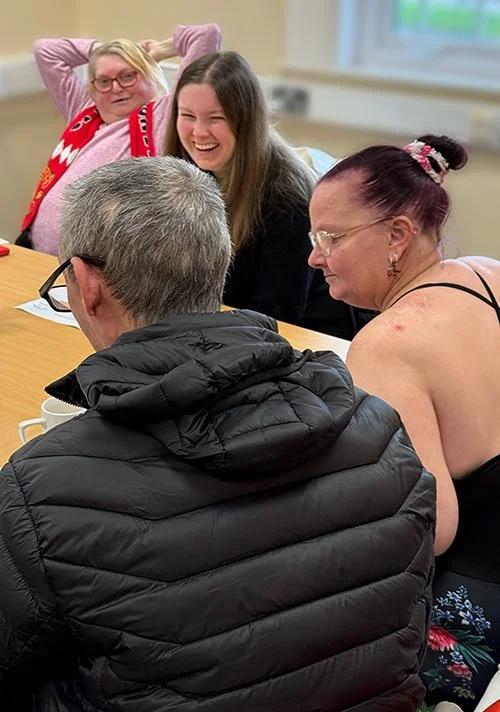 Four people sitting around a table, three women and one man, engaging in conversation and smiling, in a comfortable indoor setting.
