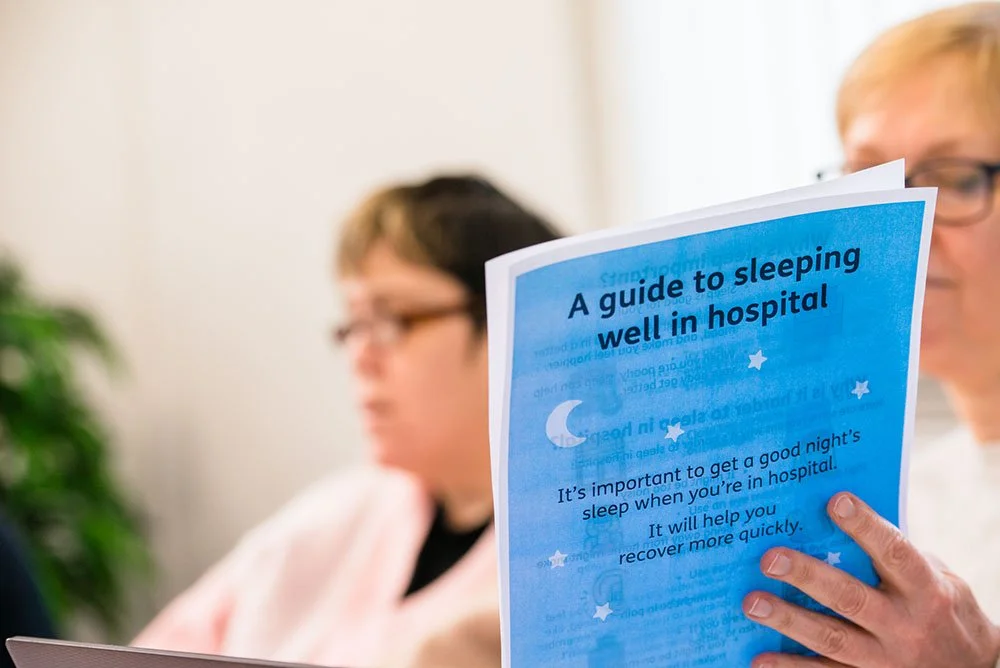 A person holding a blue booklet titled "A guide to sleeping well in hospital" with an image of a moon and stars, while other people are in the background in an office setting.