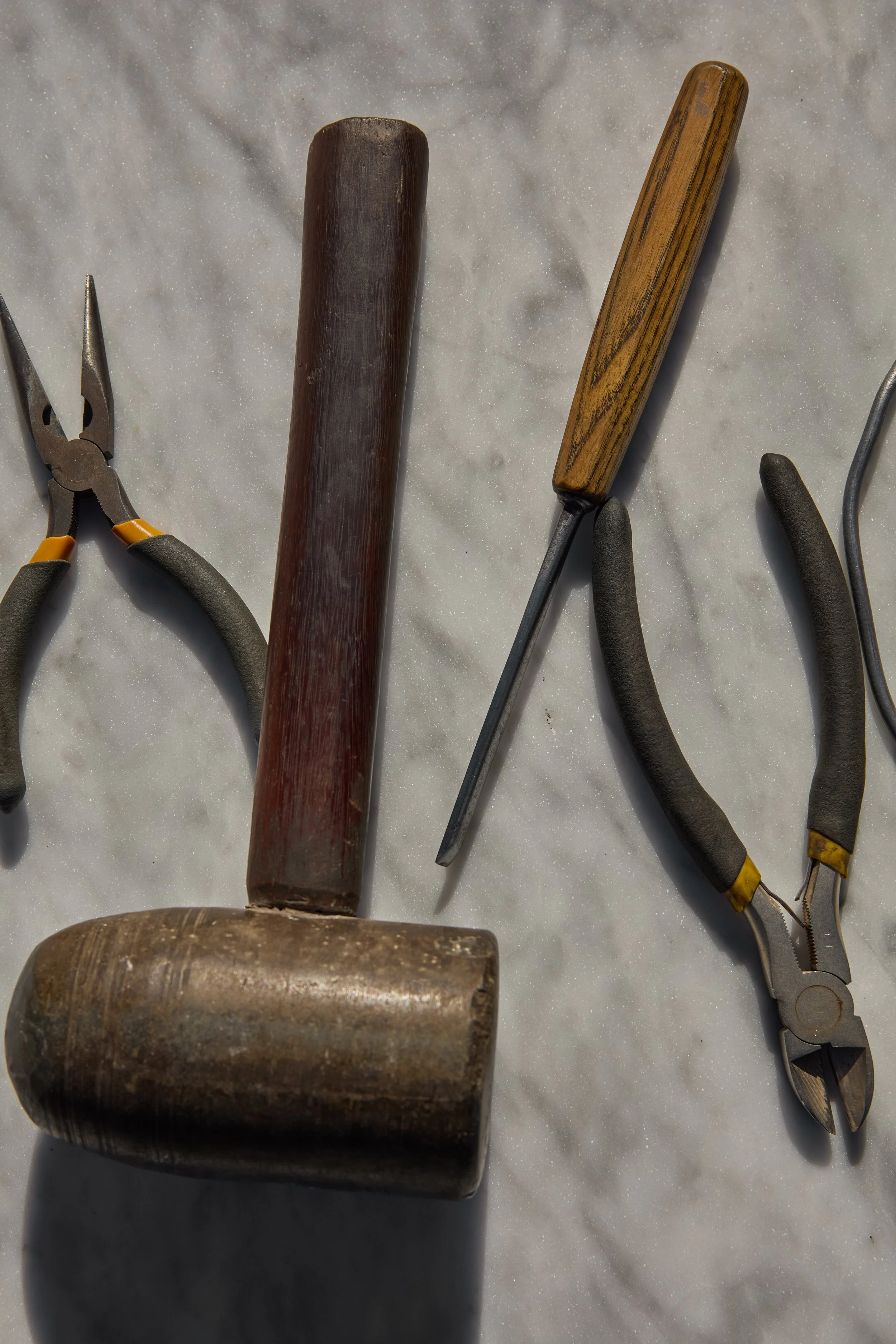 A set of jewelry-making tools including a pair of pliers, a wooden hammer, a file, and another pair of pliers, arranged on a white surface.