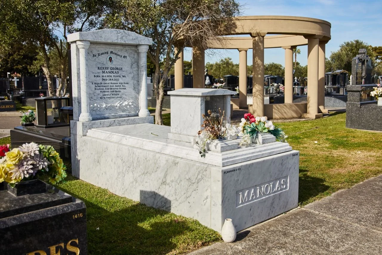 A marble grave at a cemetery with a headstone for Kerry George Manolas and a decorative circular pavilion in the background, surrounded by other gravestones and flowers.