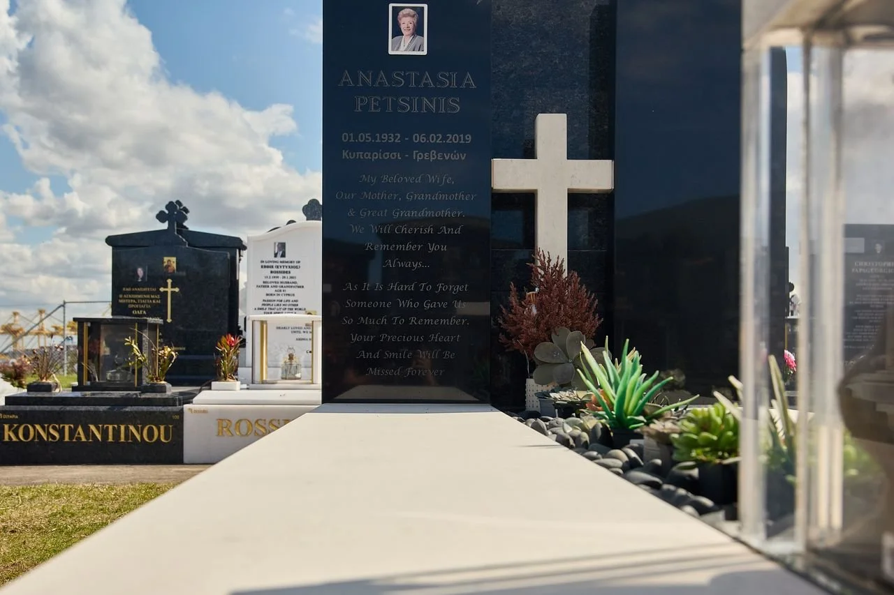 A row of gravestones and memorial plaques in a cemetery, with a focus on a black gravestone for Anastasia Petsinis, featuring a photo, name, birth and death dates, and a poem.
