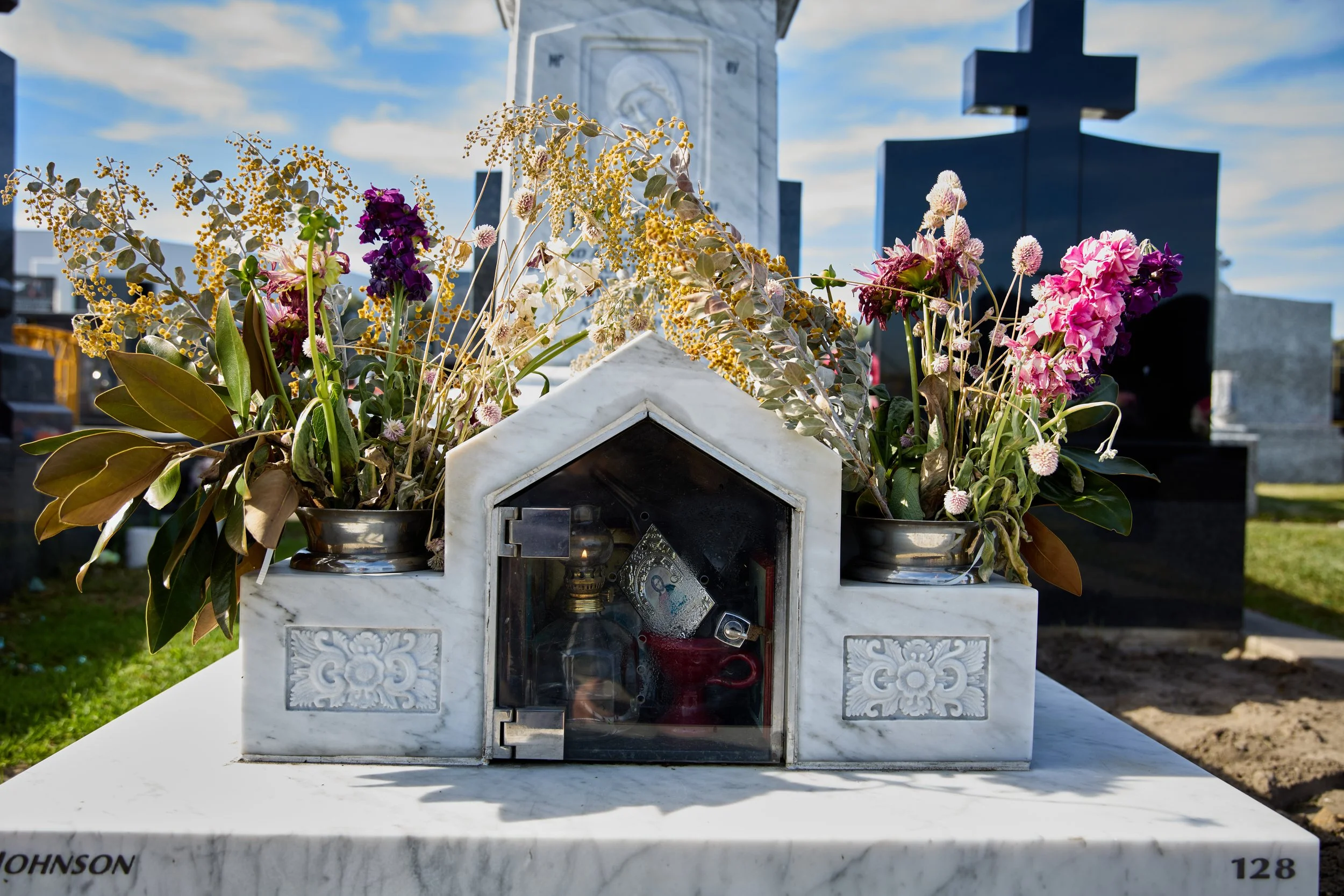 A cemetery gravestone with flowers, a small house-shaped shrine, and several other gravestones in the background.