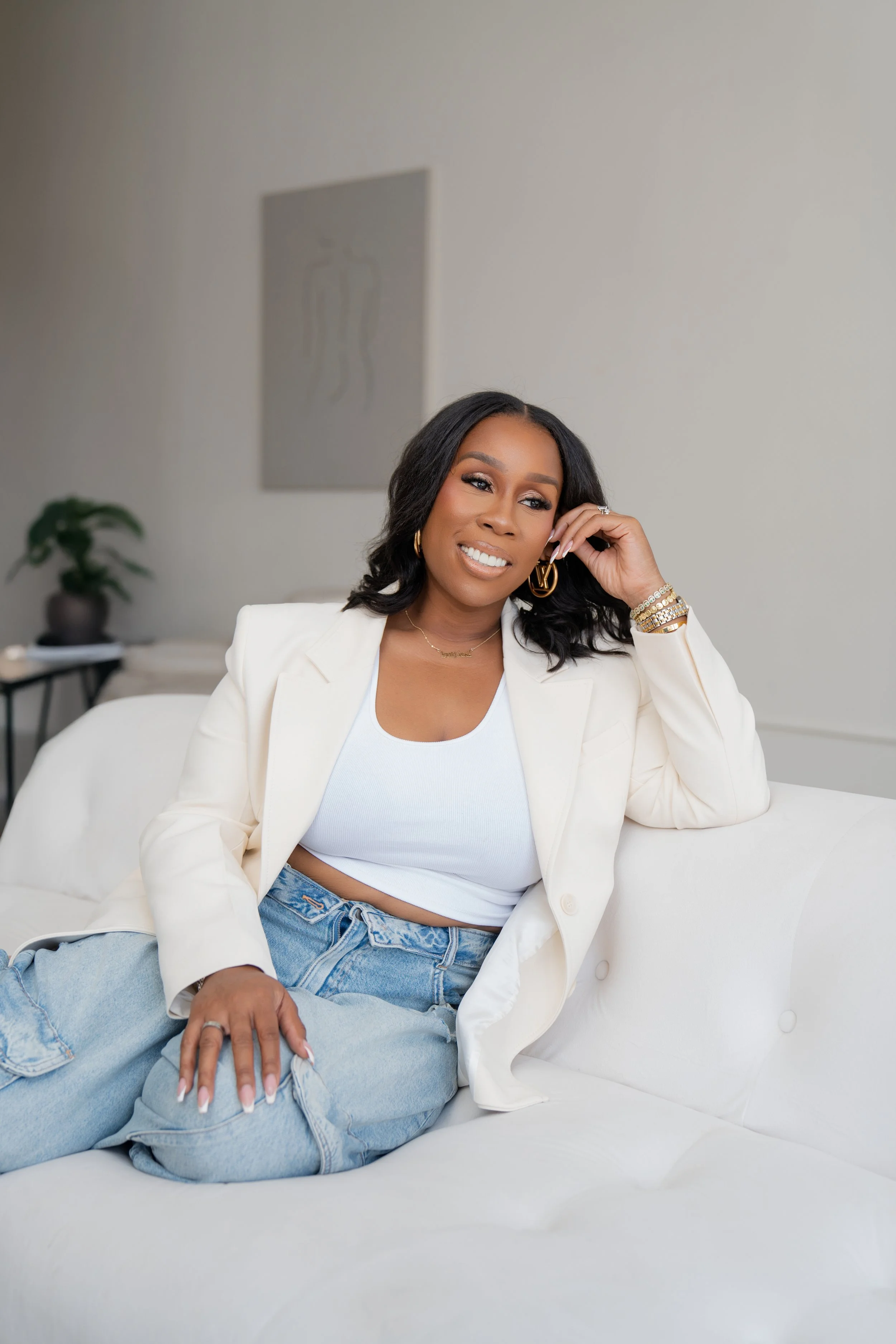 A woman sitting casually on a white sofa, smiling, wearing a white blazer, white tank top, and light blue jeans, with a potted plant, a framed art piece, and a side table in the background.