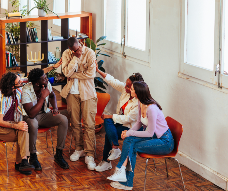 A group of five diverse young adults in a therapy or support group session in a bright room. One person appears distressed, and the others are offering comfort and support.