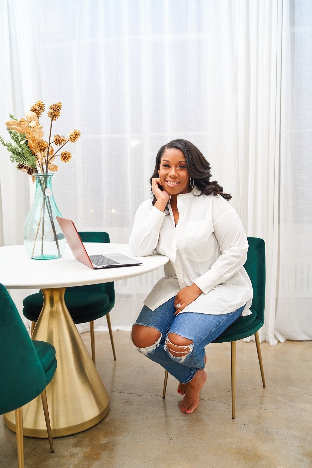 A woman cheerfully sitting at a round table with a laptop, floral arrangement, and green chairs in a bright room with white curtains.