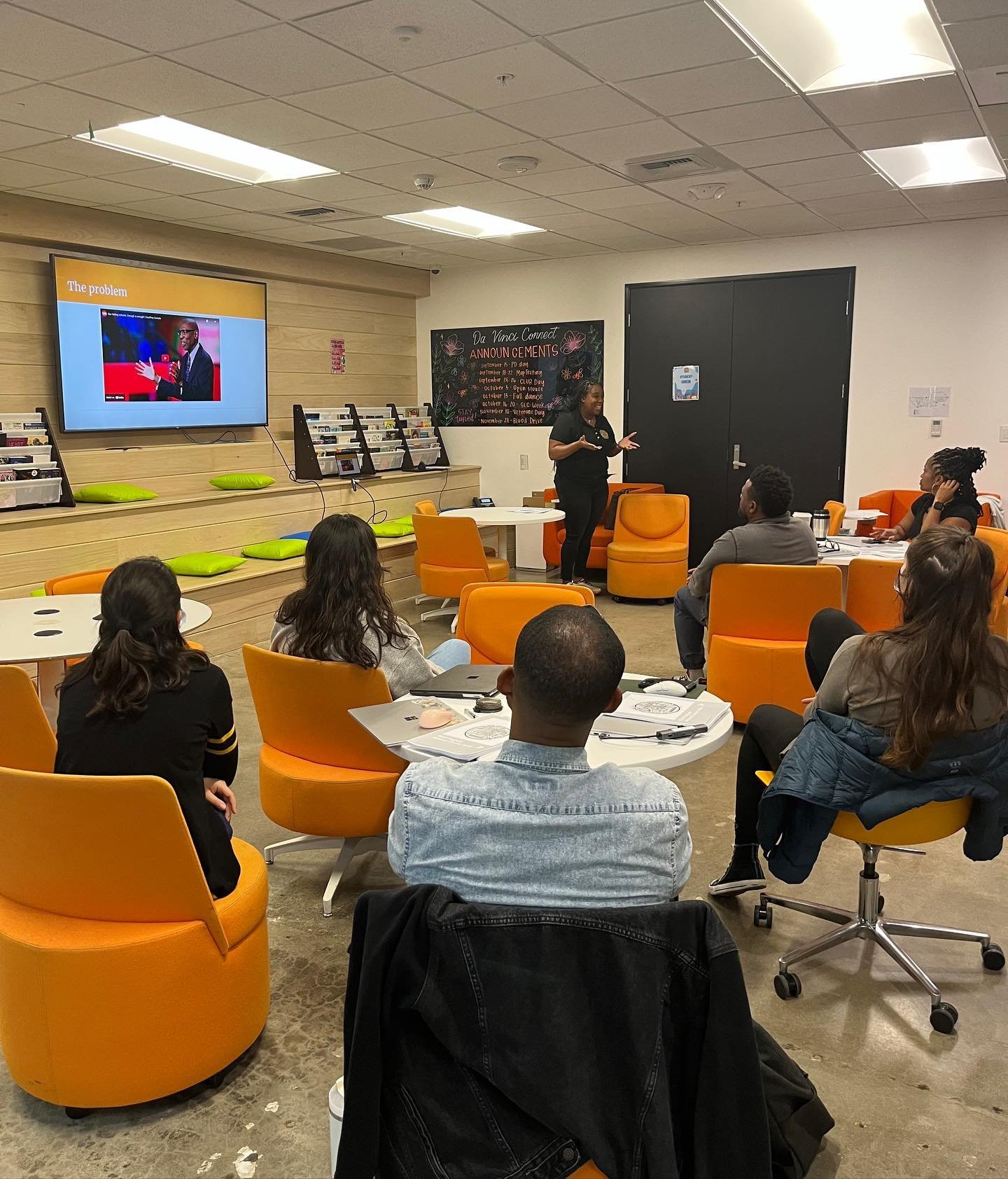 A woman is giving a presentation to a seated audience in a room with orange chairs and a large screen displaying a slide titled 'The problem' with an image of a man speaking. There is a blackboard with colorful chalk writing and a door behind the presenter.