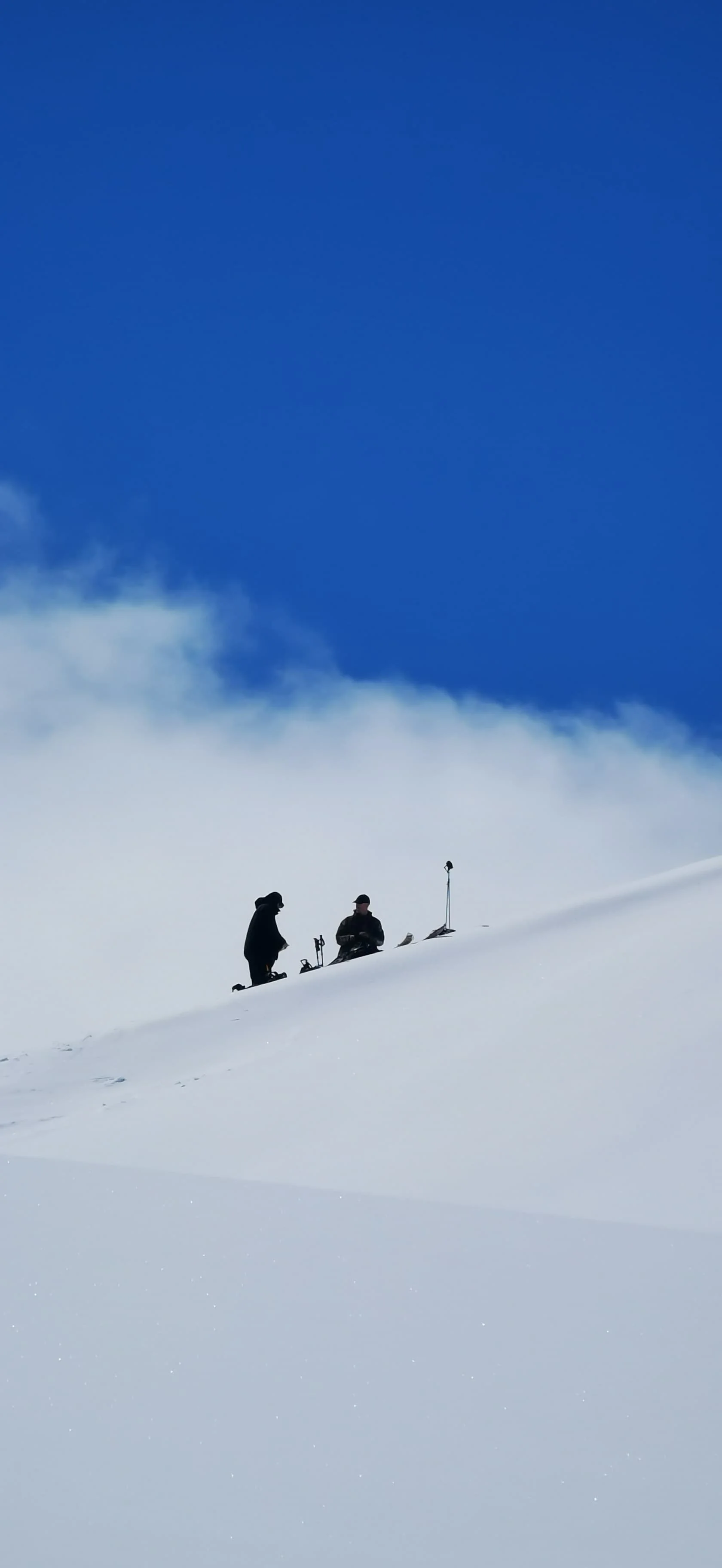 Two people on a snow-covered mountain slope, one standing and one sitting, with ski equipment, under a clear blue sky with some clouds.