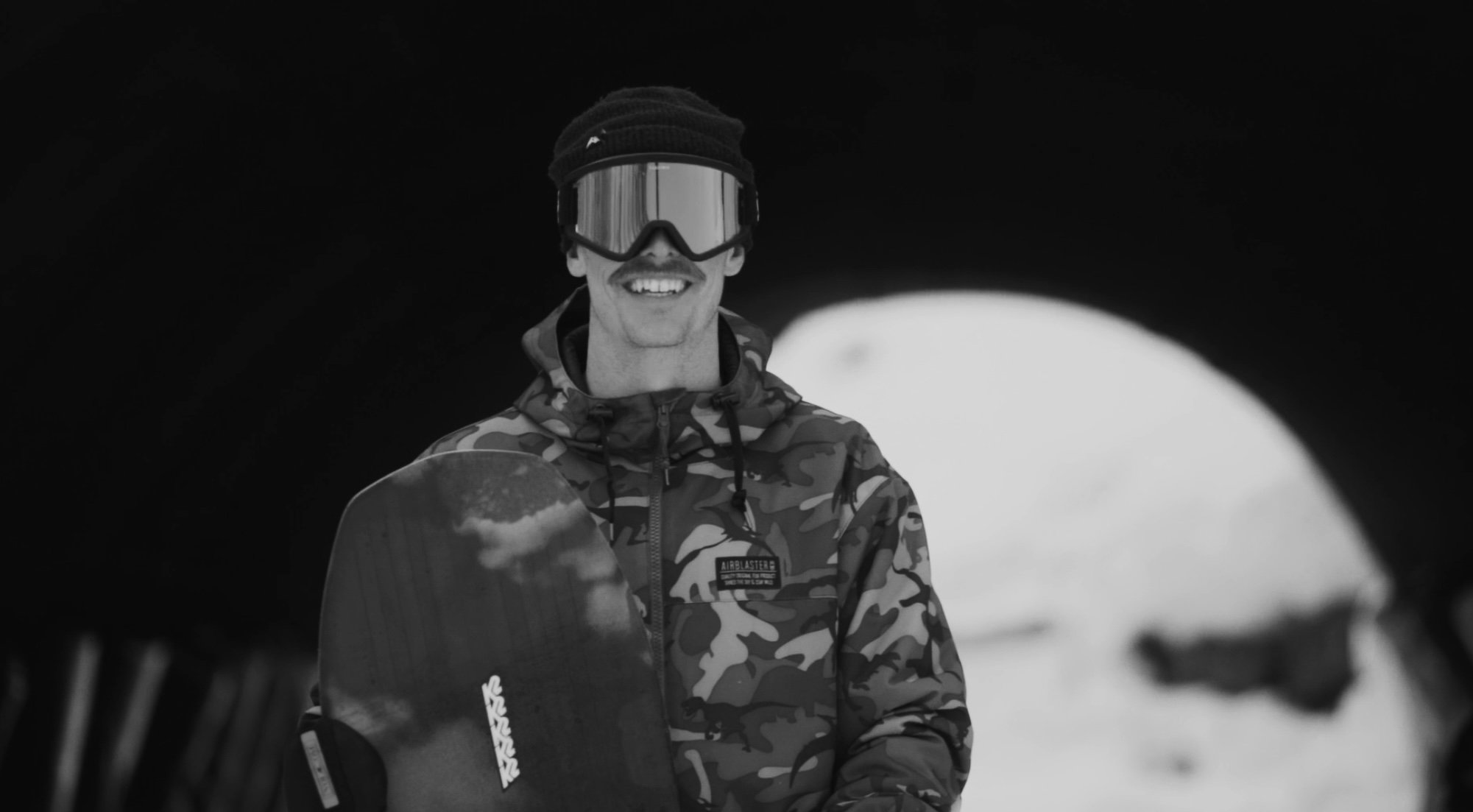 A person smiling while holding a snowboard, dressed in a camouflage jacket, wearing goggles and a beanie, standing in a snowy landscape under an archway in Tignes and Val D'isere.