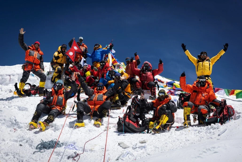 Group of trekkers or climbers celebrating on snowy mountain summit, wearing colorful winter gear and holding flags, with prayer flags and blue sky in background.
