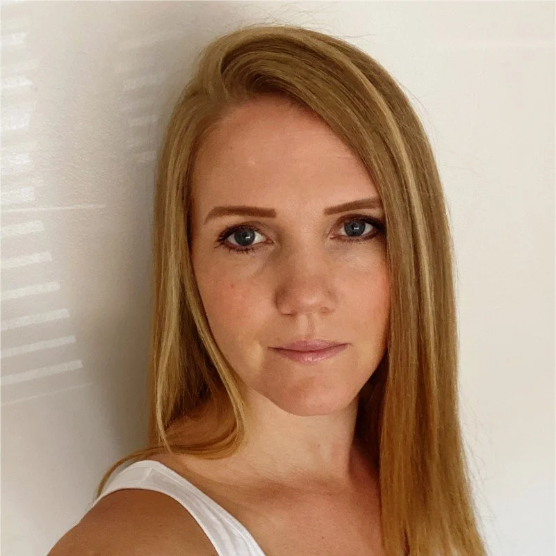 A woman with long red hair, blue eyes, and light makeup looking at the camera in front of a plain light-colored wall.