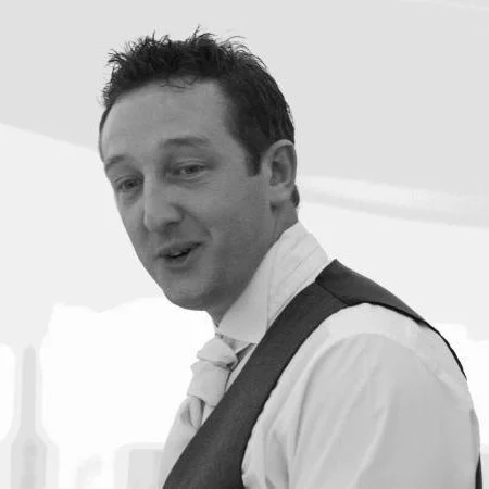Black and white photo of a man with short dark hair, wearing a white shirt with a tie and dark vest, looking slightly to the side.