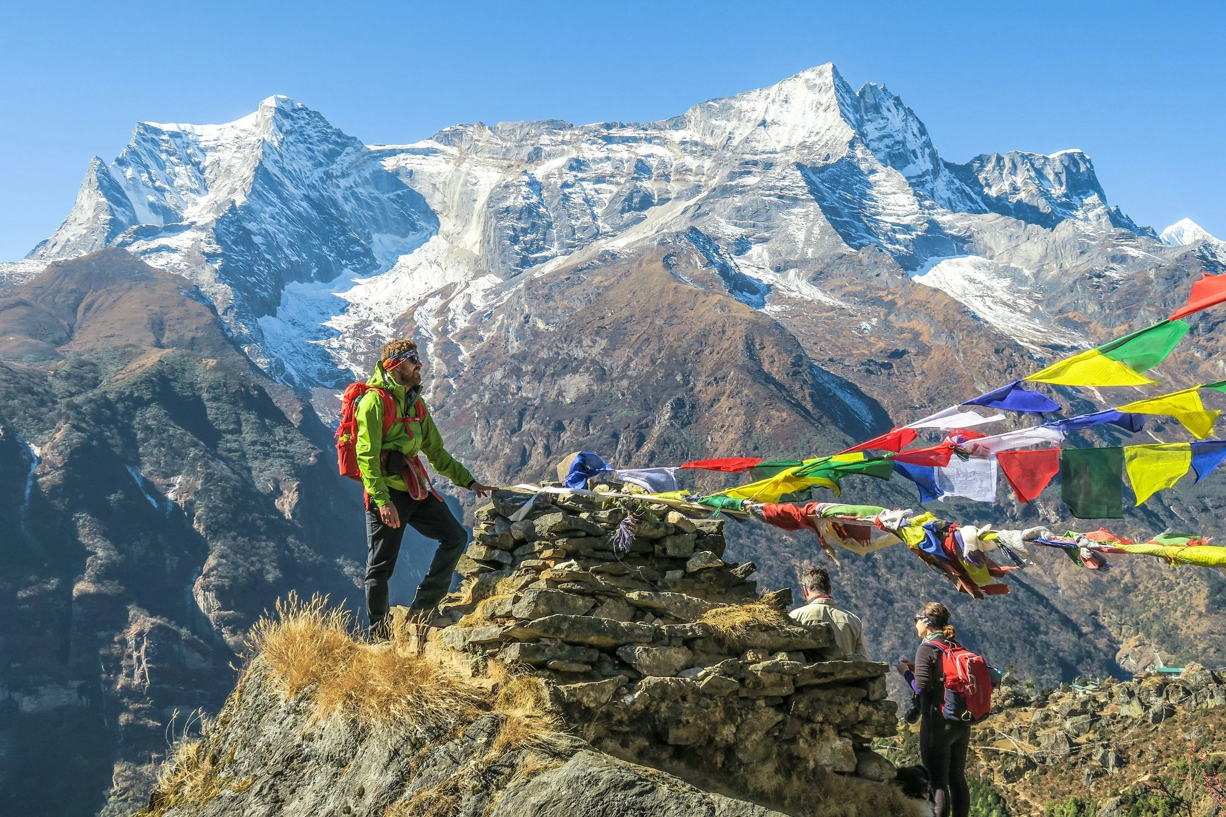 Hikers in a mountainous region with snow-capped peaks, colorful prayer flags, and rocky terrain.