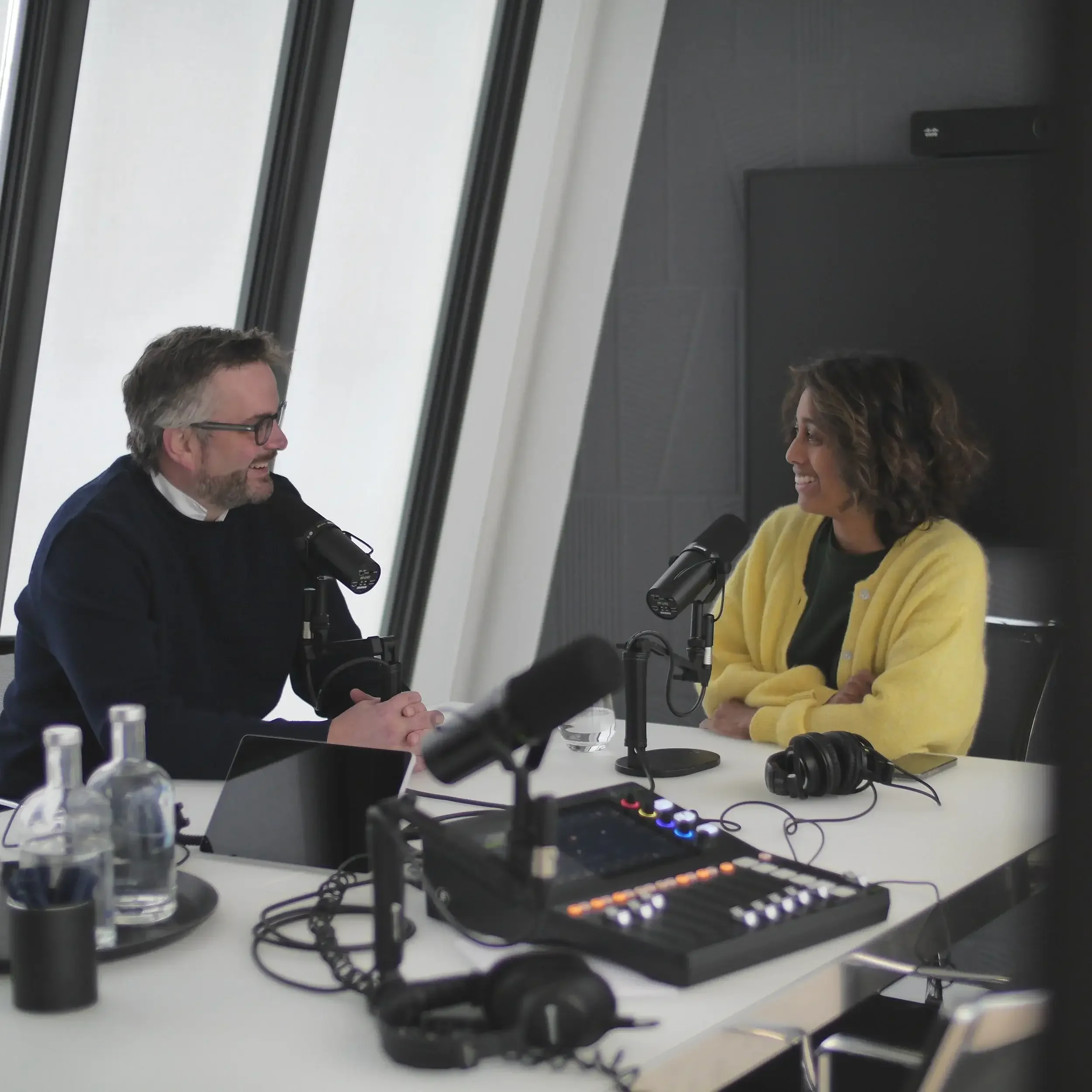 Deux personnes discutant dans une salle de conférence, avec un équipement audio et un ordinateur portable visibles sur la table.