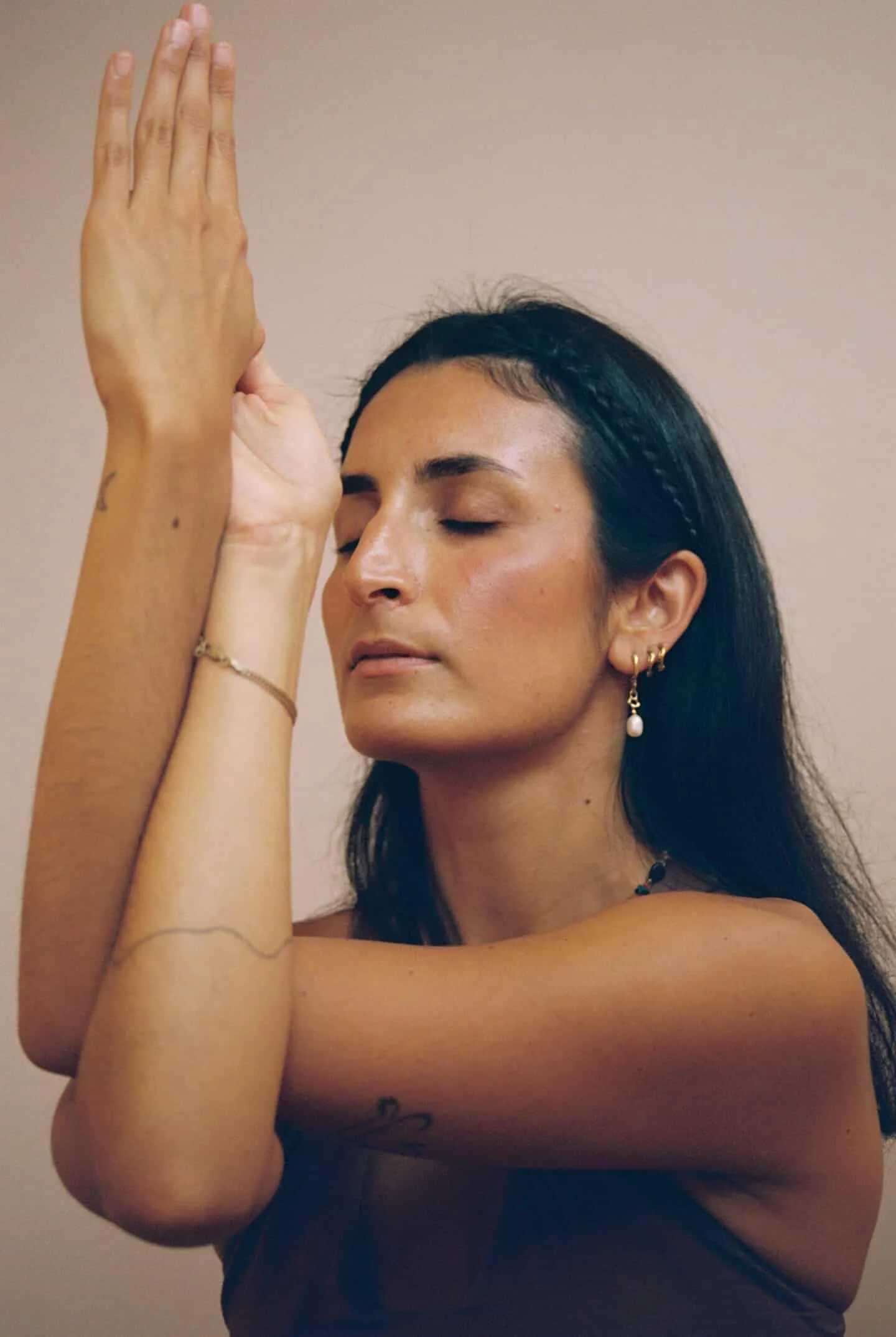 A woman with dark hair, wearing earrings and a necklace, has her eyes closed and is holding her hands together above her head in a prayer or meditation pose, against a plain beige background.