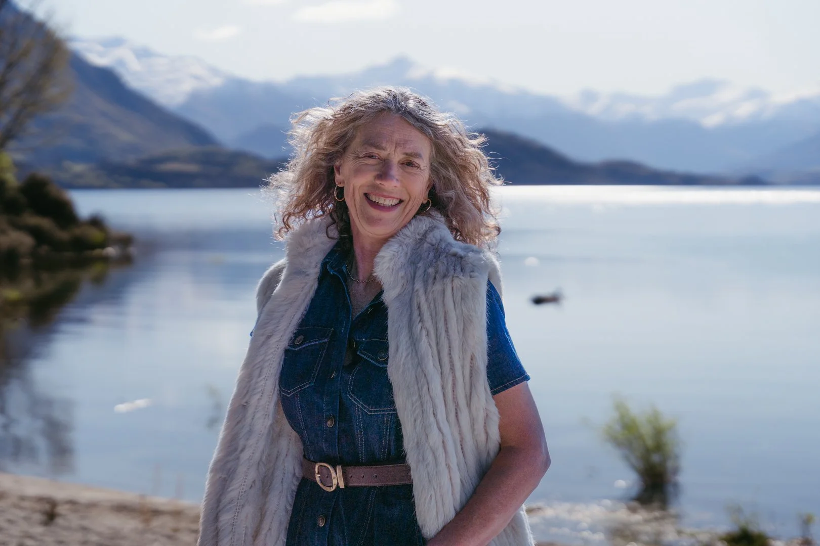 A smiling woman with curly gray hair wearing a denim shirt and a fur vest stands near a lakeshore, with mountains in the background under a partly cloudy sky.