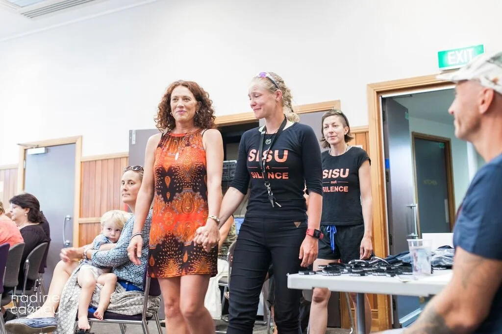 Women, including one wearing a black T-shirt with red text, stand together in a room during an event. There are other people sitting and a table with items on it in the background.