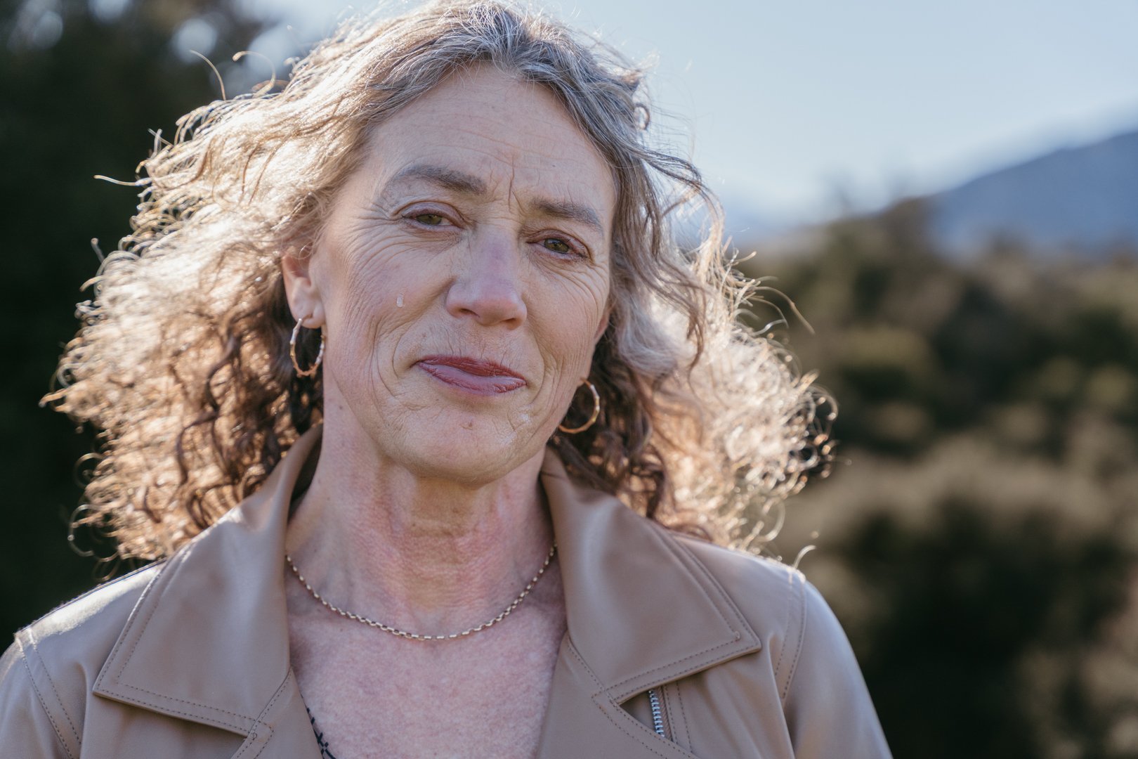 Close-up of a middle-aged woman with curly gray hair, wearing a beige jacket and gold jewelry, outdoors with a blurred natural background.