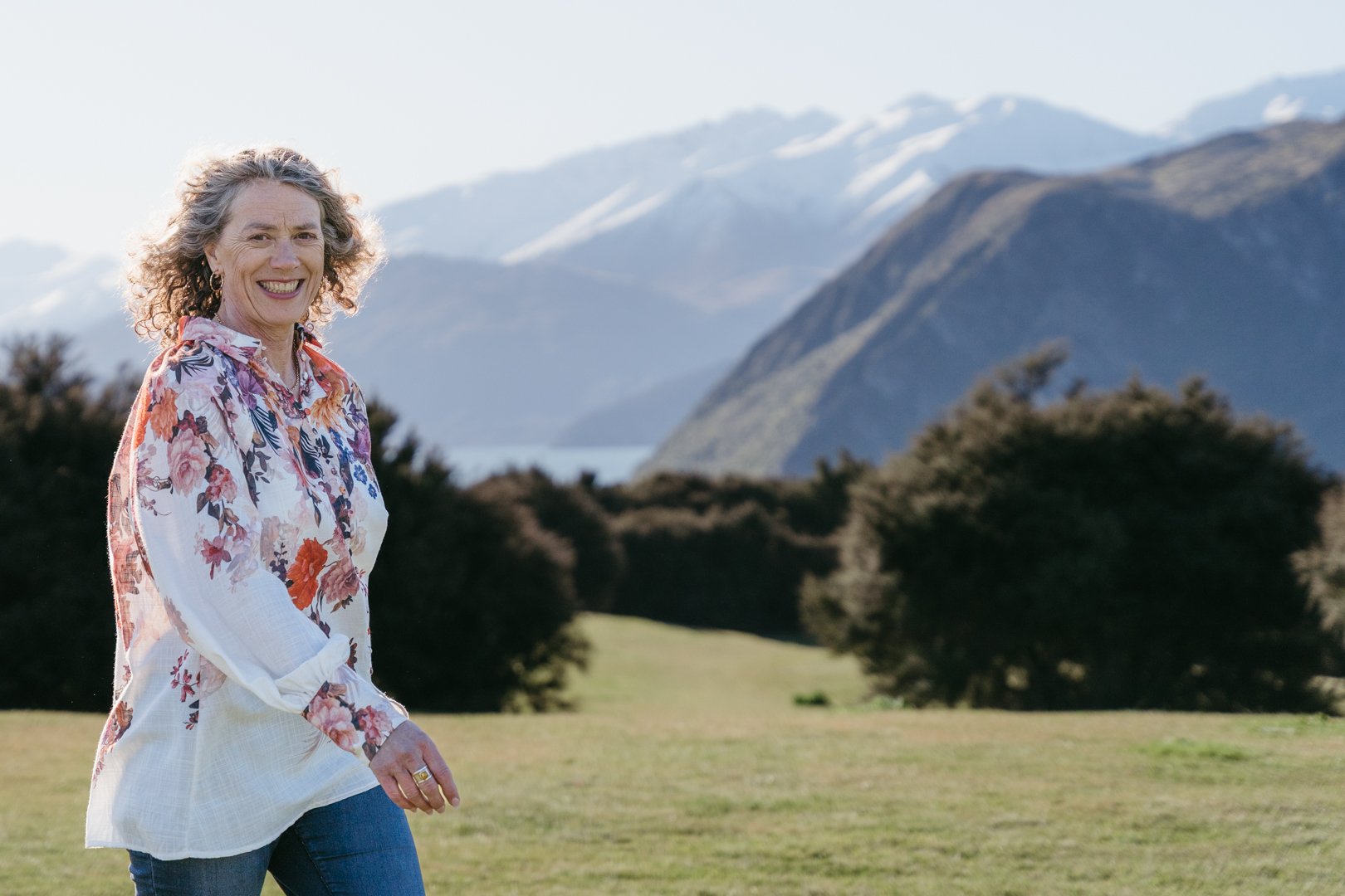 A woman smiling outdoors in a grassy area with mountains and a body of water in the background, wearing a floral blouse and jeans.