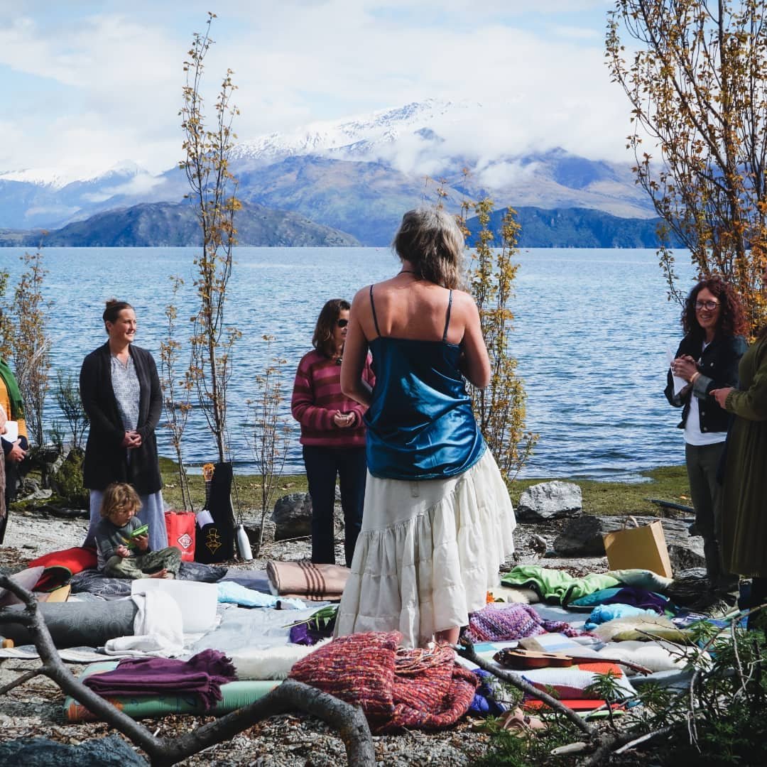 Group of women and children standing on rocks near a lake with snow-capped mountains in the background. Some are smiling, and items like blankets and backpacks are on the ground.