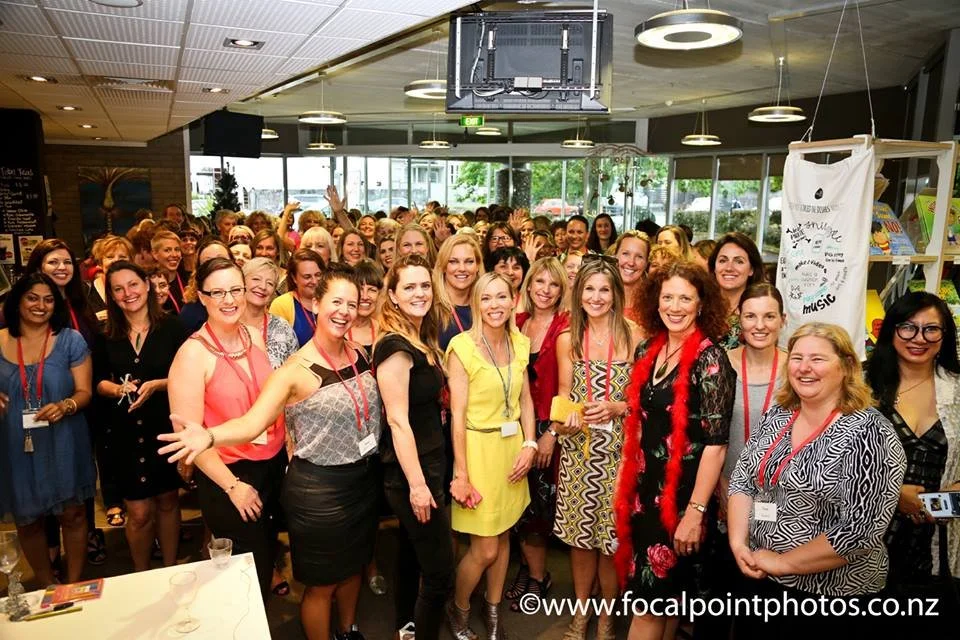 Group of smiling women gathered in a well-lit indoor space, posing for a photo at what appears to be a social or professional event.
