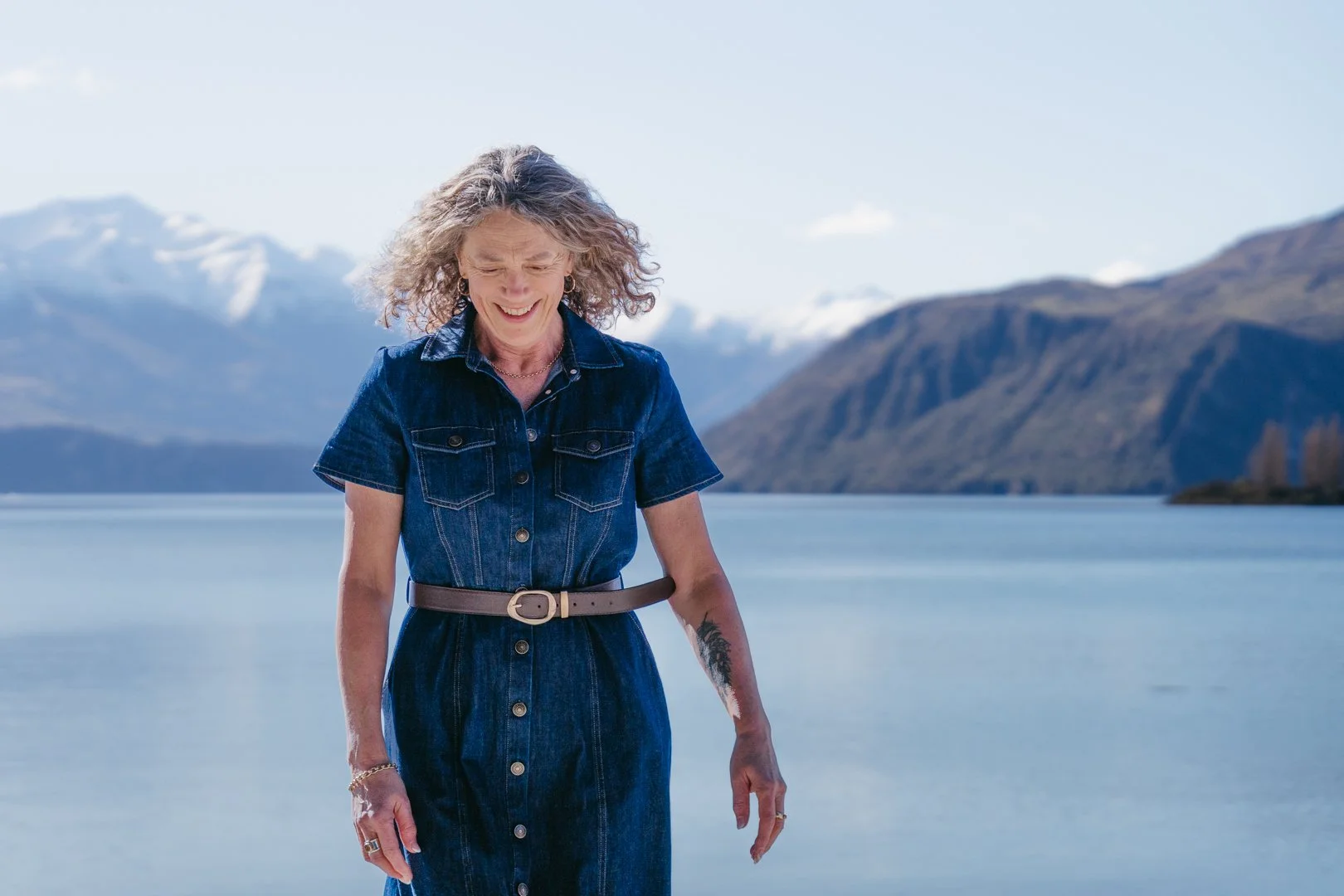 A woman in a denim dress standing near a lake with mountains in the background, smiling and looking down.