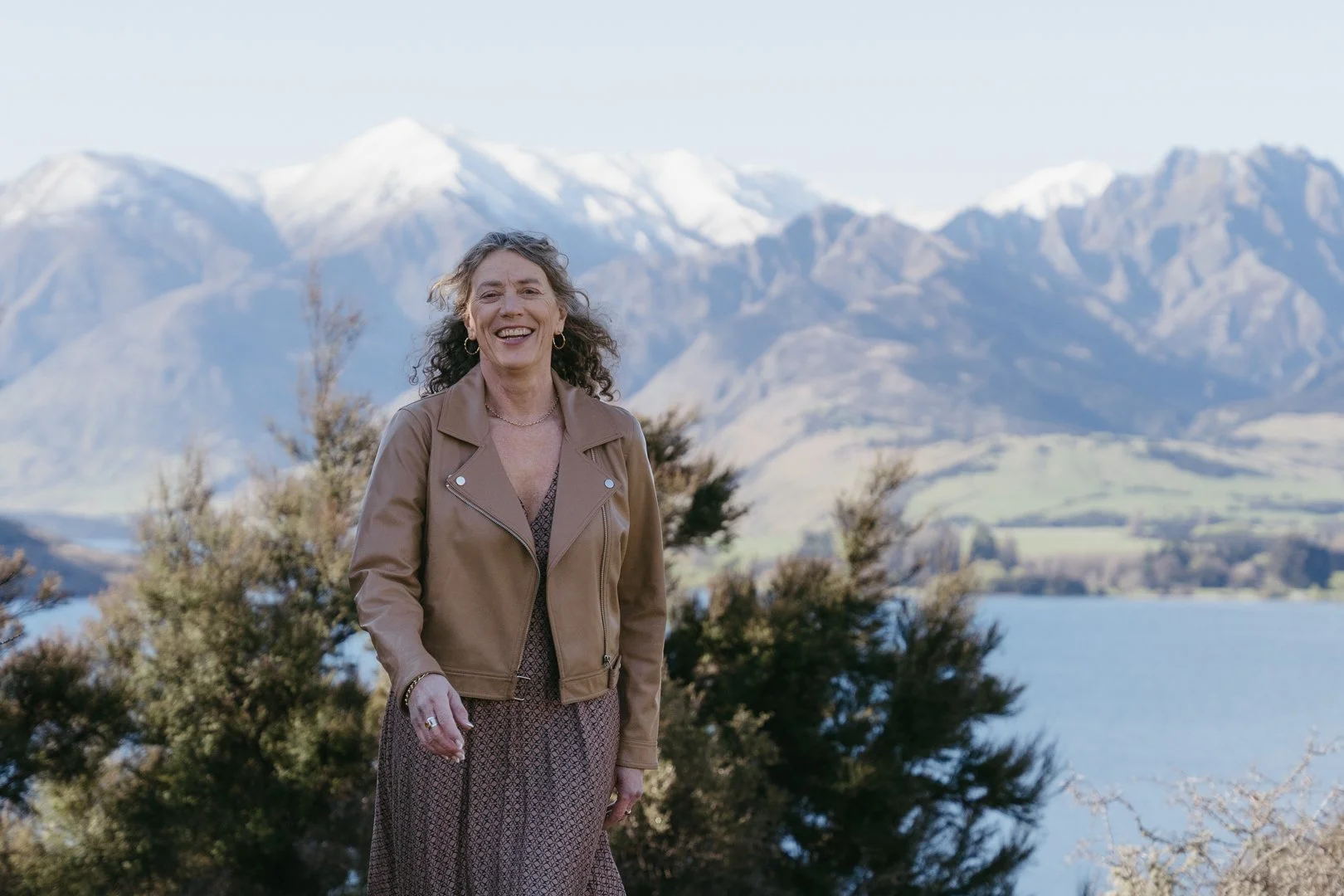 A smiling woman with curly hair, wearing a brown leather jacket and a patterned dress, standing outdoors near a lake with snow-capped mountains in the background.