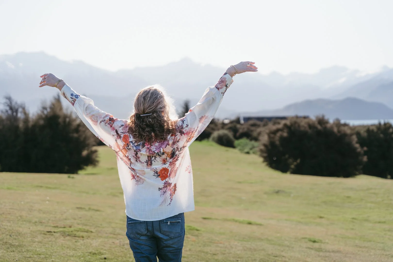Older woman with curly hair wearing a floral blouse and jeans with arms raised in a grassy field with trees and mountains in the distance.