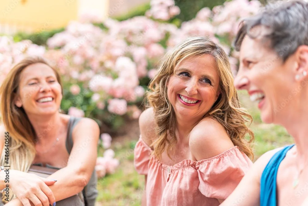 Three women laughing and smiling outdoors with pink flowers in the background.