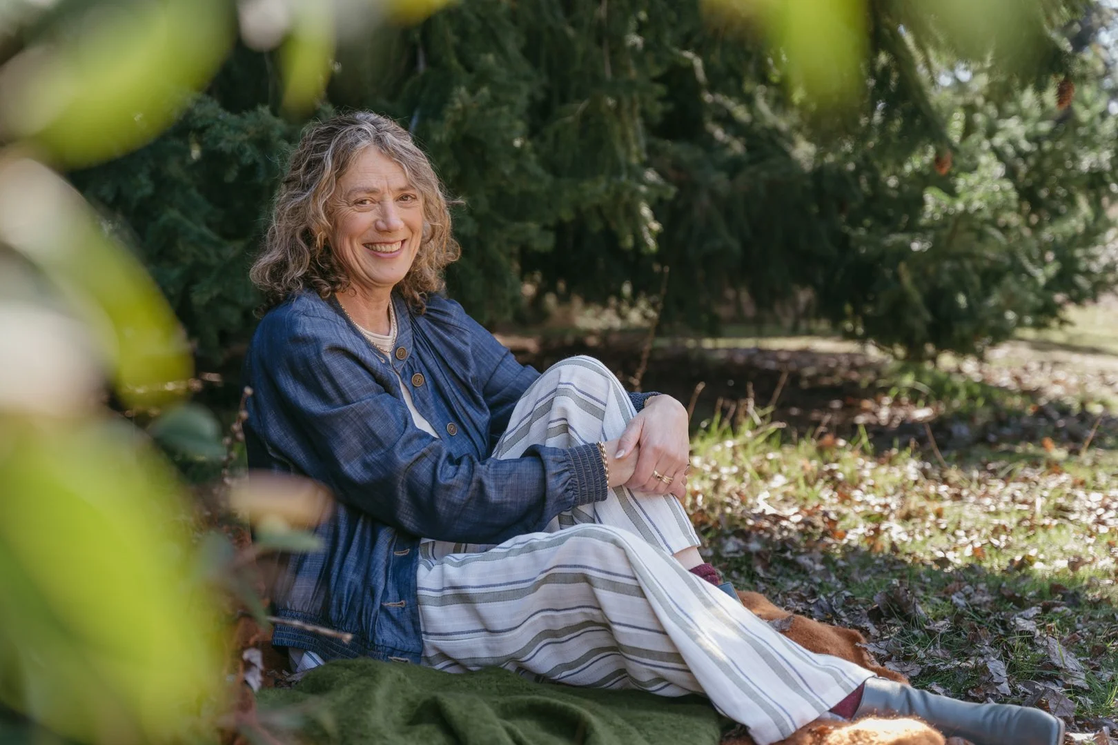 A smiling woman with curly hair sitting outdoors on a green blanket amongst trees and fallen leaves.